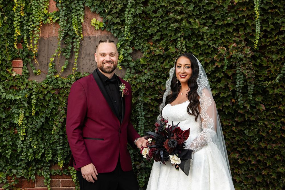 Smiling bride in a white lace gown and veil holds a dark red bouquet beside a groom in a burgundy velvet jacket before an ivy-covered wall.