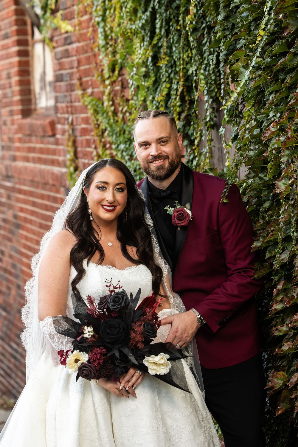 Smiling bride in a lace-veiled white gown holding a dark floral bouquet beside a groom in a burgundy jacket near an ivy-covered brick wall.