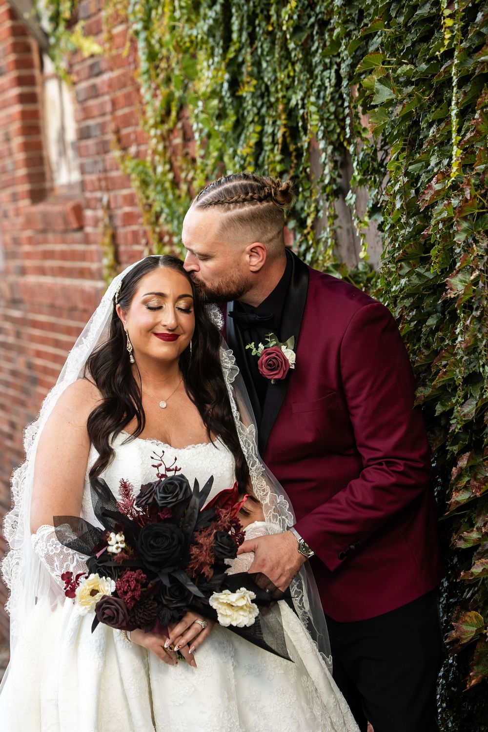 Groom in burgundy jacket kisses bride's forehead as she holds a dark floral bouquet beside an ivy-covered brick wall.