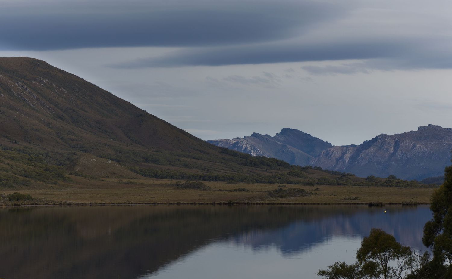 Calm mountain lake reflecting rugged peaks and grassy hillsides under a soft, cloudy sky in a remote wilderness landscape.