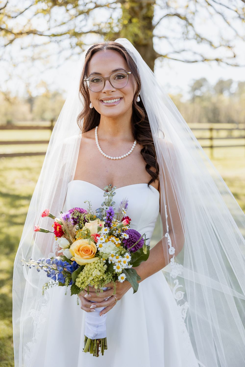Smiling bride in glasses, pearl necklace and long veil holds a colorful wildflower bouquet while standing outdoors in a sunny field.