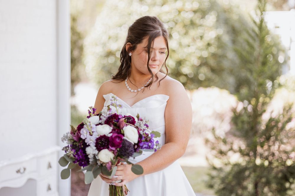 Bride in a white strapless gown and pearl necklace holds a lush purple, white and burgundy bridal bouquet outdoors on her wedding day.
