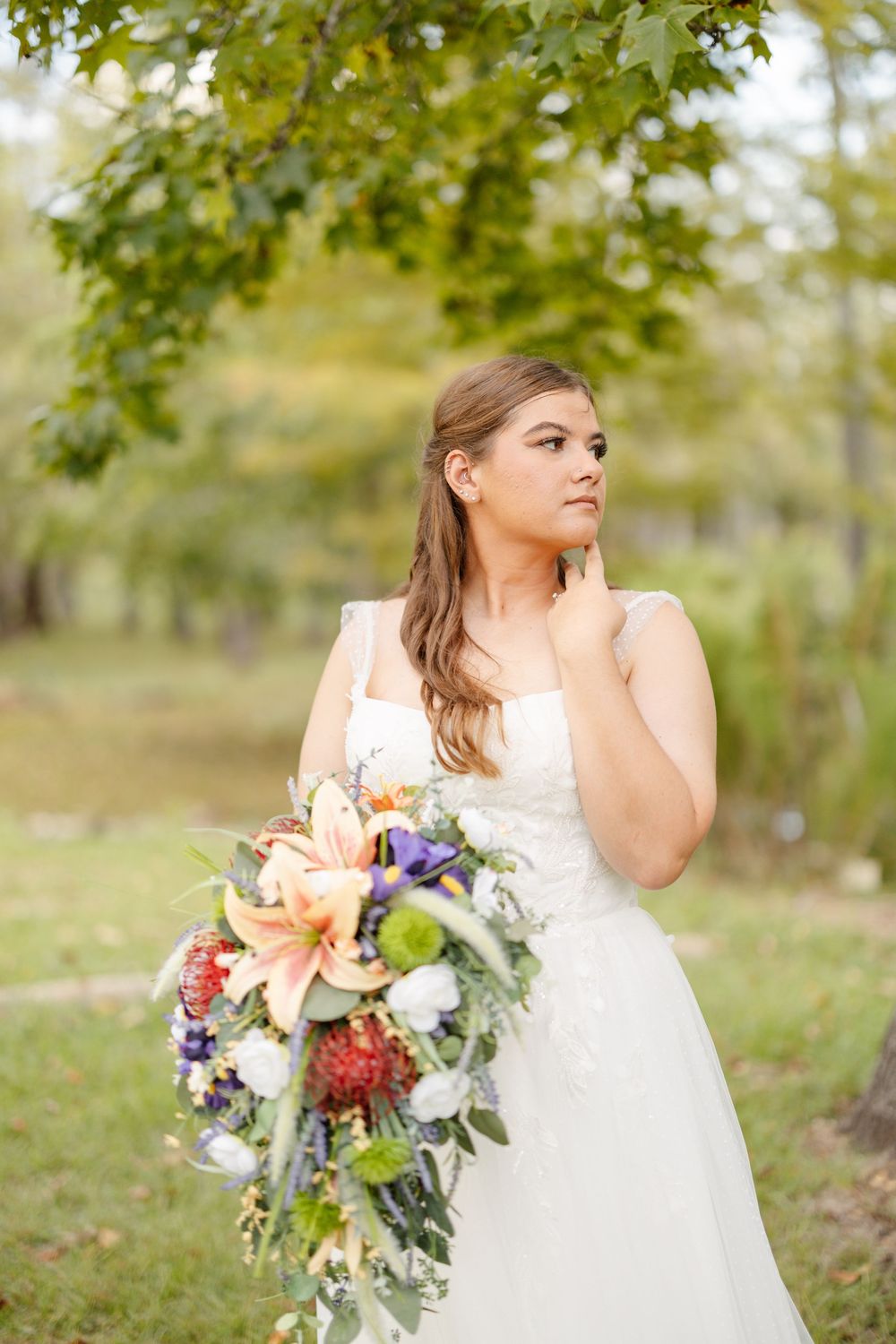 Bride in a white lace gown holding a cascading bouquet of orange lilies, purple and white flowers while posing outdoors under green trees.