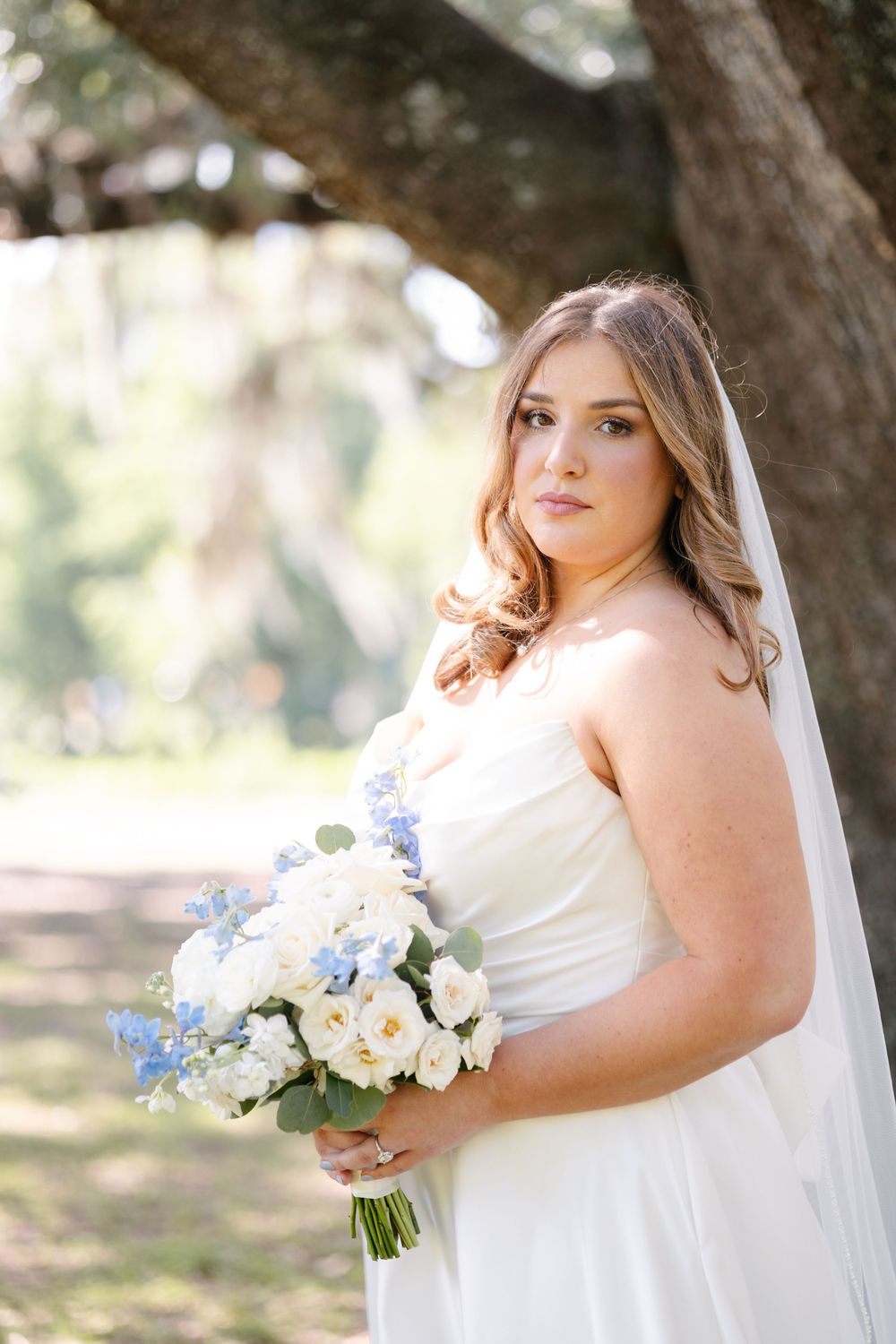 Bride in a white strapless gown and long veil holds a bouquet of white and blue flowers beneath a large oak tree.
