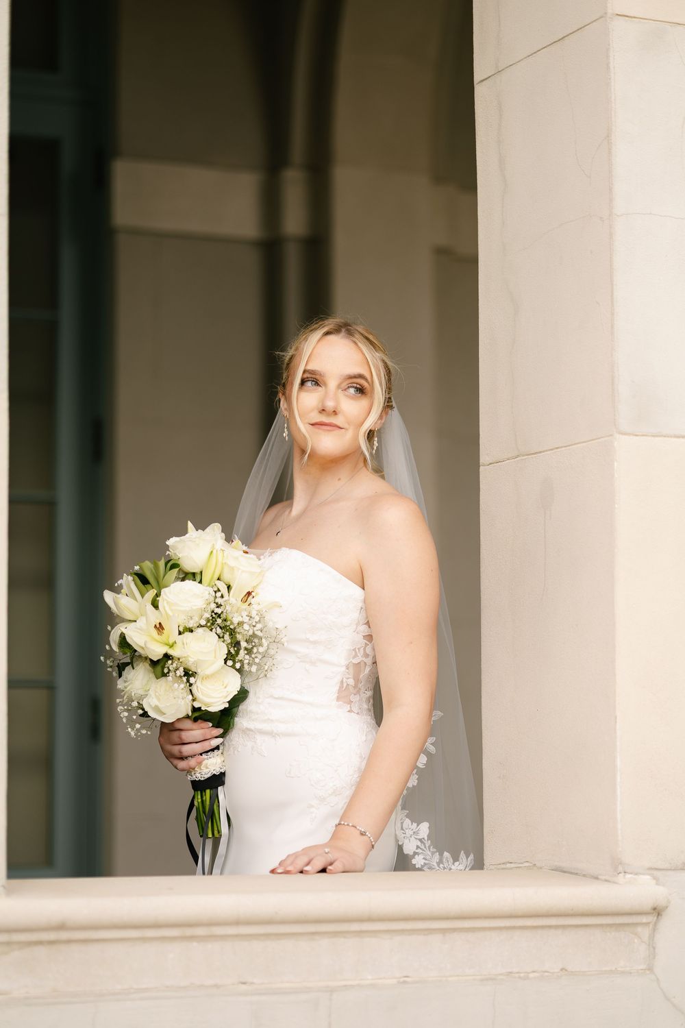 Bride in a strapless white lace gown and veil holds a white calla lily bouquet while gazing out from a stone window ledge.