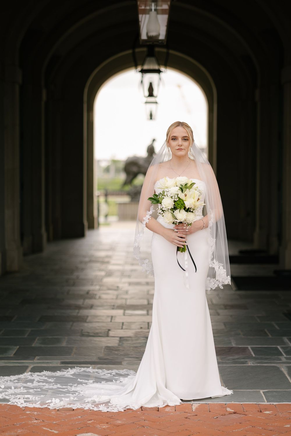 Bride in an elegant fitted white gown with lace veil holding a white floral bouquet while standing in a stone archway corridor.