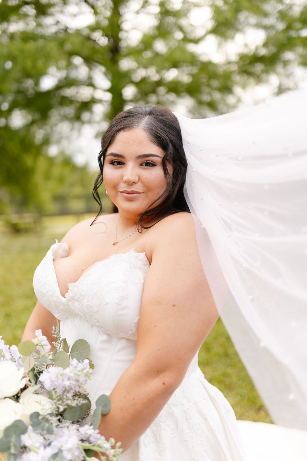Smiling bride in a white strapless lace wedding gown holds a bouquet of white and lavender flowers as her veil blows in the breeze outdoors.