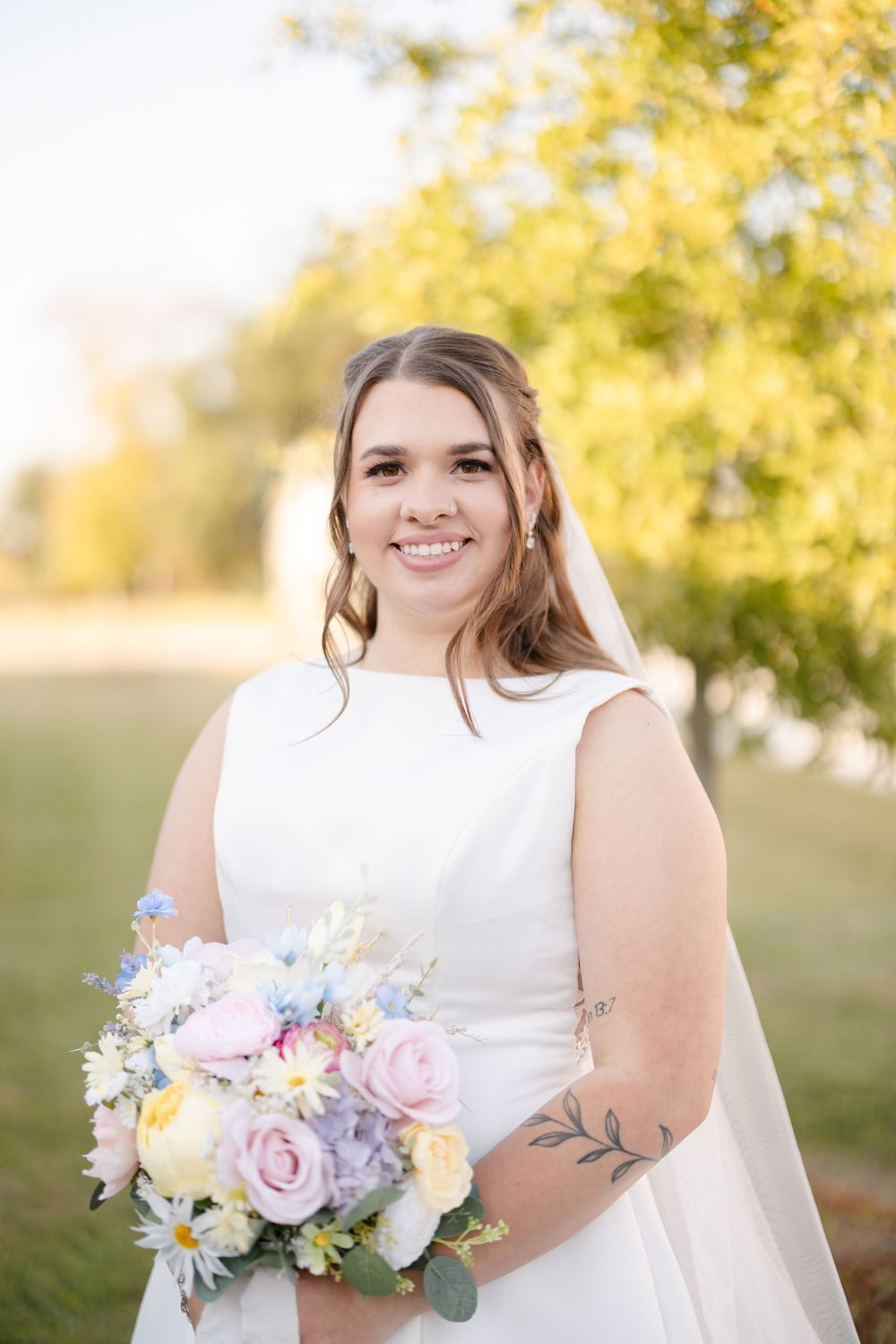 Smiling bride in a white sleeveless wedding gown and veil holds a pastel bouquet of pink, blue and yellow flowers outdoors.