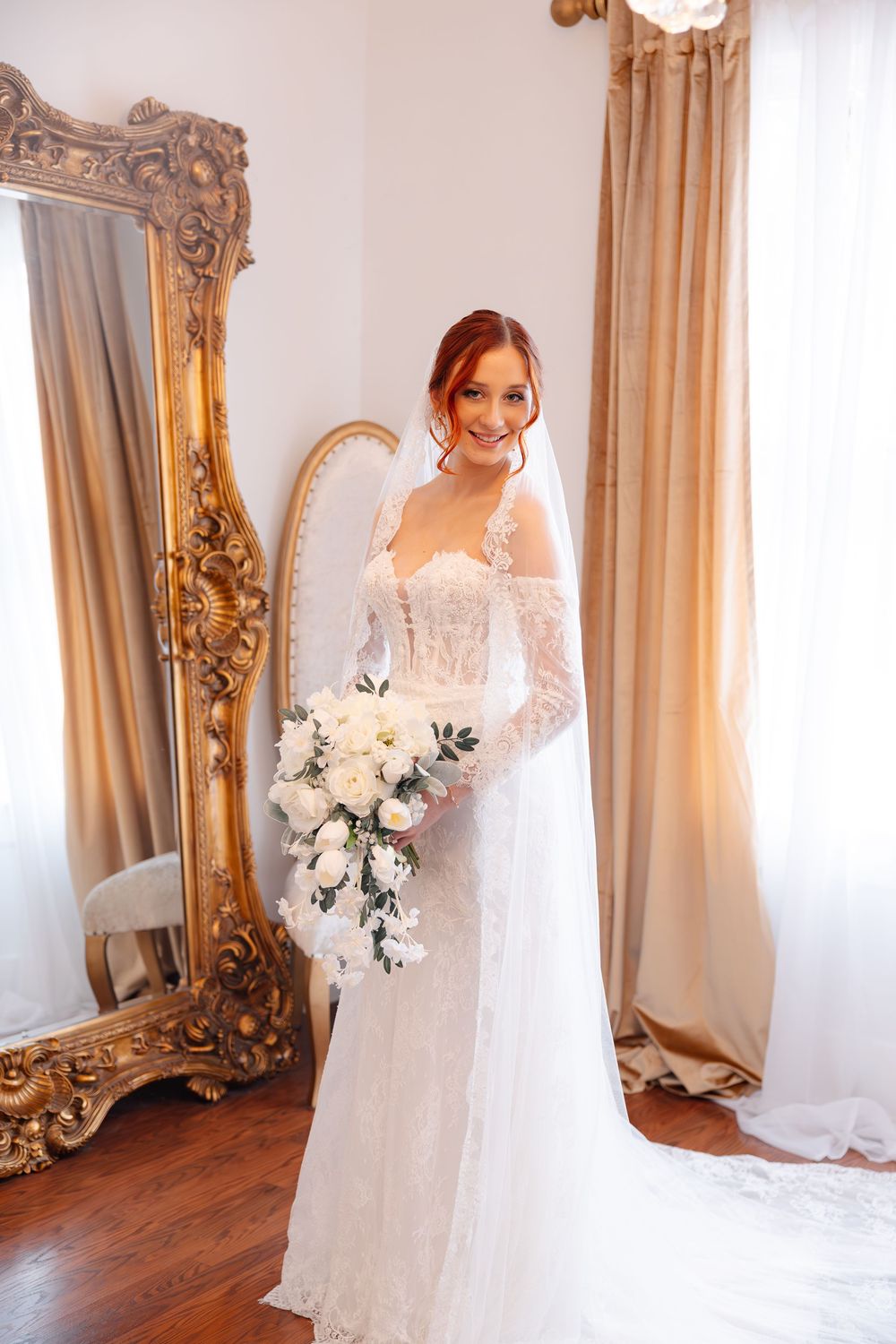 Smiling bride in a lace wedding gown and long veil holding a white rose cascade bouquet beside an ornate gold mirror.