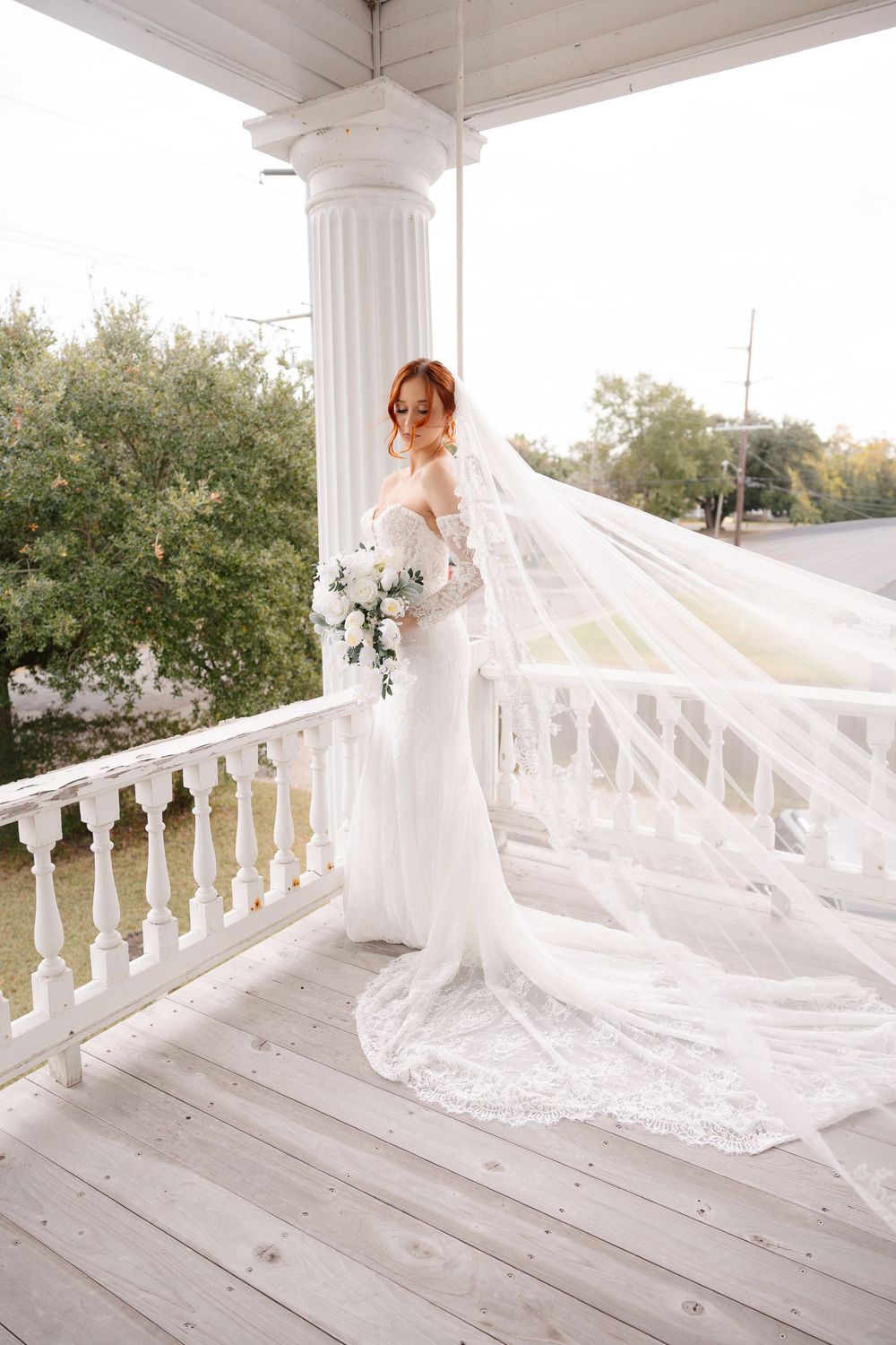 Bride in an off-the-shoulder lace wedding gown with a long flowing veil holds a white bouquet on a white porch beside a tall column.