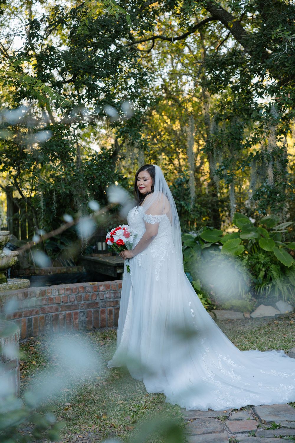 Bride in a white off-the-shoulder lace wedding gown and long veil holding a red and white bouquet in a lush garden beside a brick fountain.