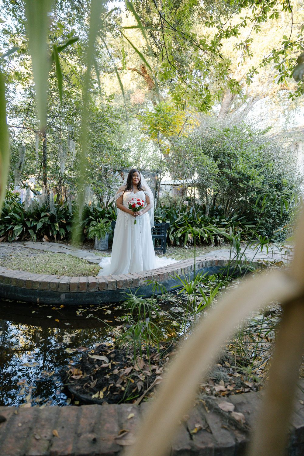 Bride in a white gown holding a red and pink bouquet stands beside a brick-edged garden pond surrounded by lush greenery.