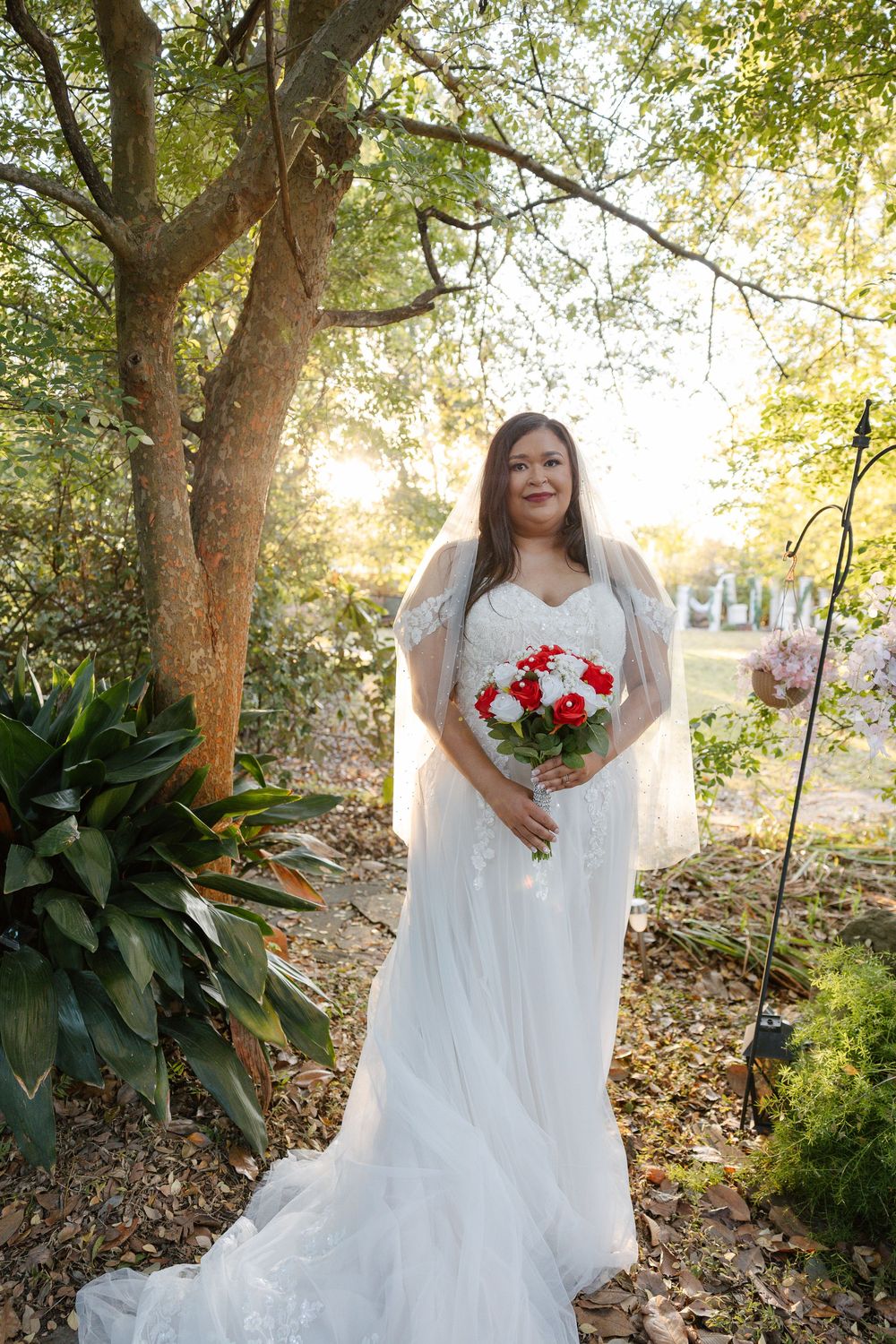 Bride in a white lace wedding gown and veil holds a red and white rose bouquet while standing outdoors beneath sunlit trees.
