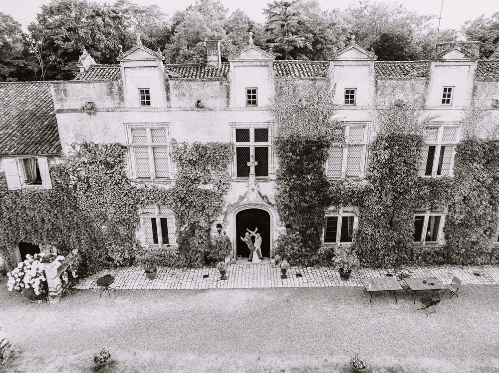 Vue aérienne noir et blanc d'un château en pierre couvert de lierre, avec son entrée en arche et son allée de gravier
