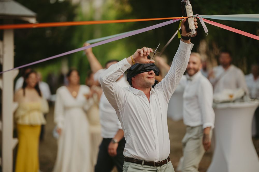 Jeu de mariage : homme aux yeux bandés tirant sur des rubans colorés pendant la fête en extérieur