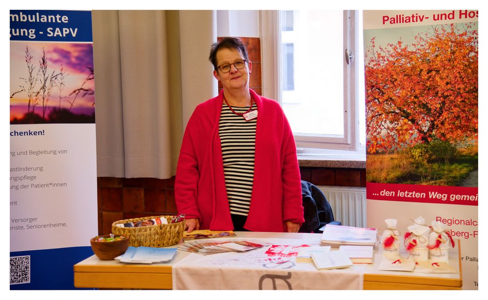 Frau in roter Strickjacke steht an einem Informationsstand zum Thema Palliativmedizin auf einer Gesundheitsmesse.