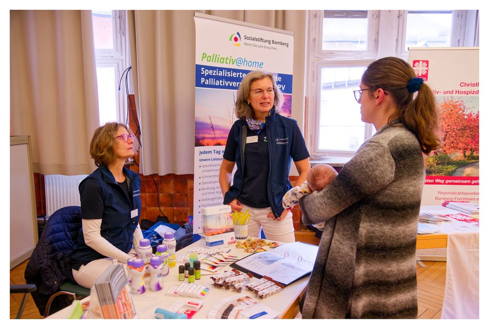 Zwei ehrenamtliche Helfer im Gesundheitswesen unterhalten sich auf einer Messe mit einem Besucher über Palliativpflege zu Hause.