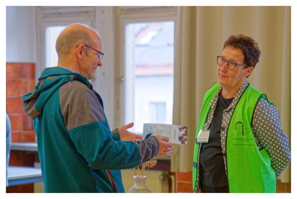 Ein Mann in einer türkisfarbenen Jacke unterhält sich mit einem Freiwilligen in einer grünen Weste bei einer Gemeinschaftsveranstaltung in e