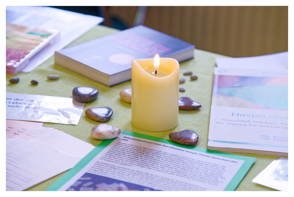 Eine brennende Kerze, umgeben von glatten Steinen und aufgeschlagenen Büchern auf einem grünen Tisch.