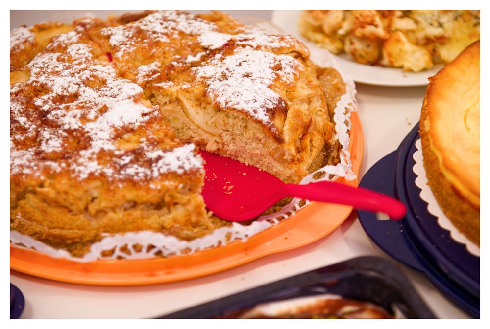 Ein mit Puderzucker bestäubter Apfelkuchen mit einem roten Spatel auf einem orangefarbenen Teller.