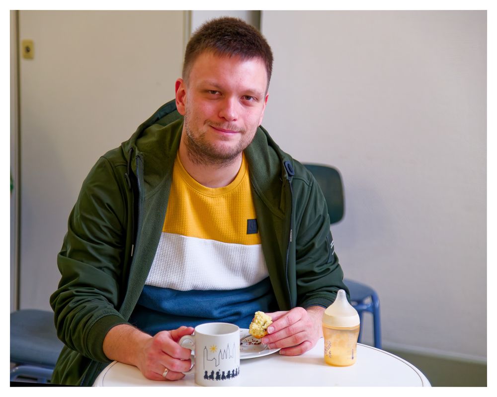 Ein lächelnder junger Mann genießt Kaffee und Kuchen an einem kleinen weißen Tisch.