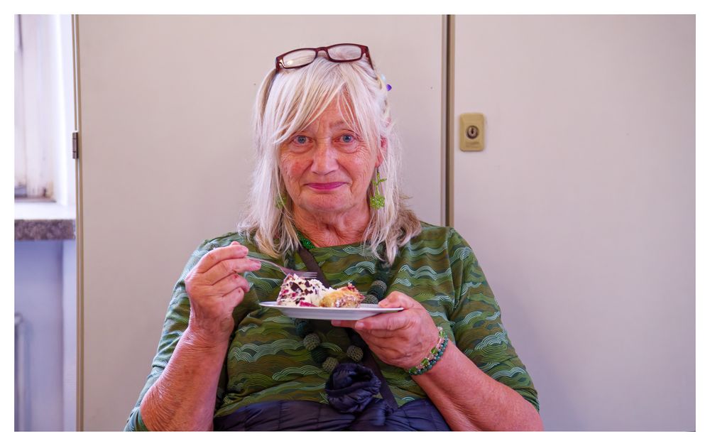 Eine ältere Frau mit weißem Haar hält einen Teller mit Essen und lächelt.