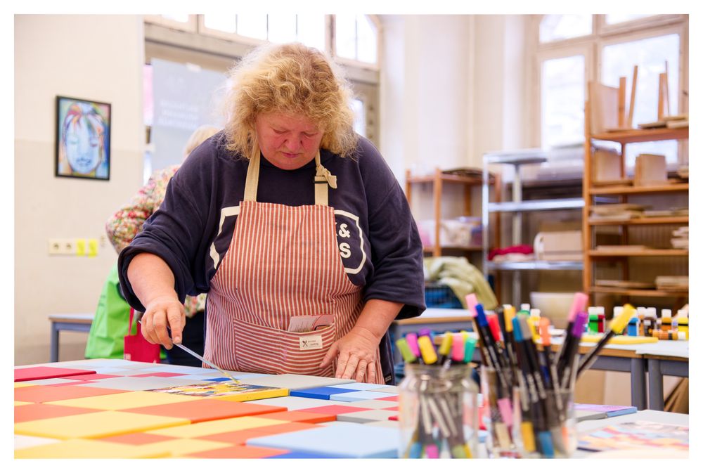 Eine Frau mit lockigem Haar und einer Schürze schneidet an einem Basteltisch buntes Papier.