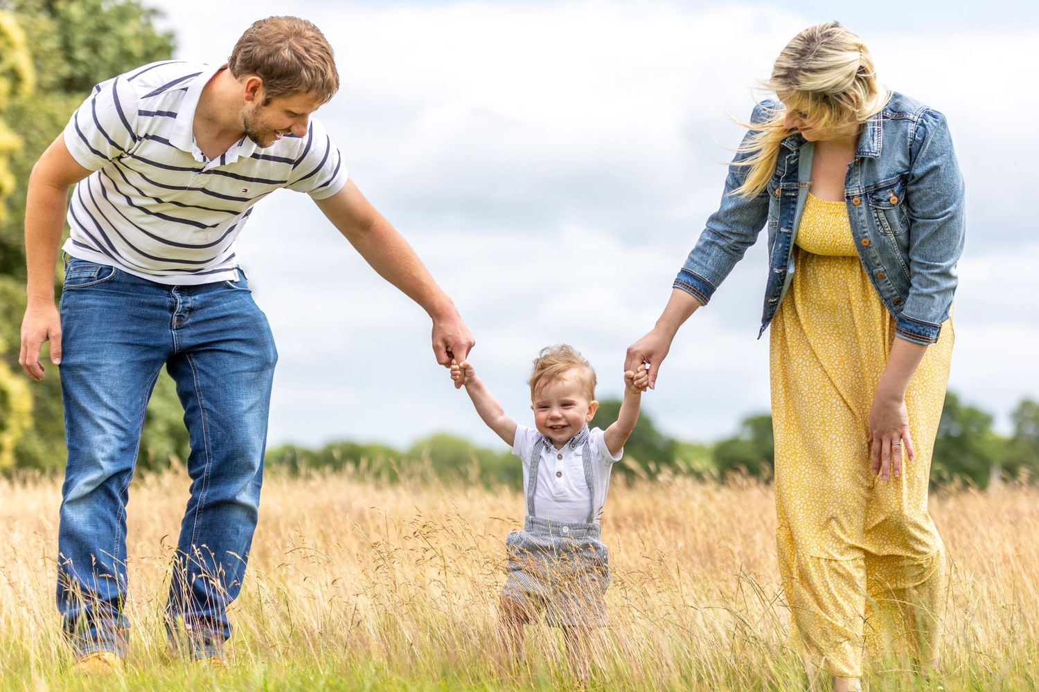 Happy family with parents holding toddler son's hands while walking in a golden meadow on a sunny day.