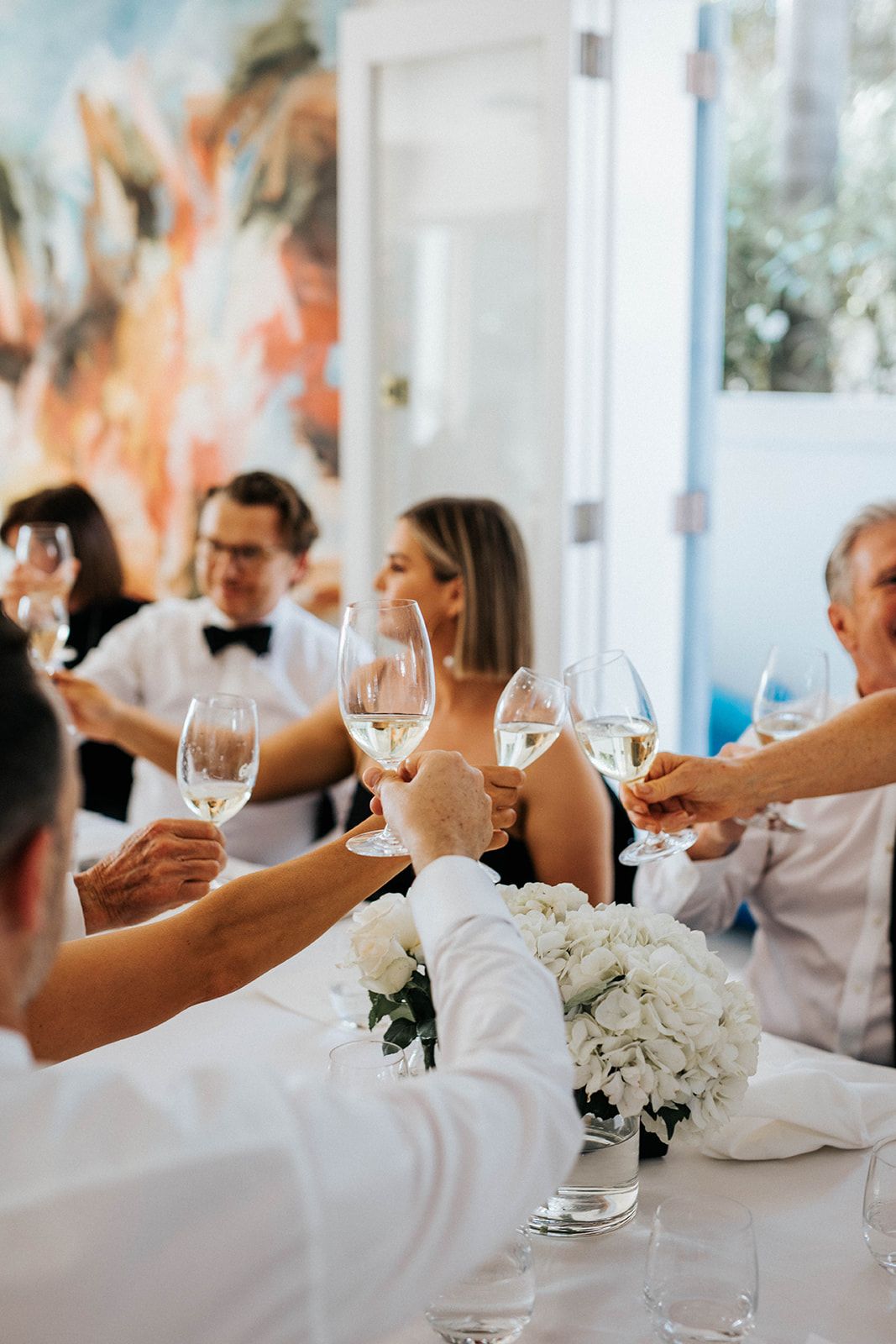 Guests raising white wine glasses in a toast at an elegant wedding reception with white floral centerpieces.