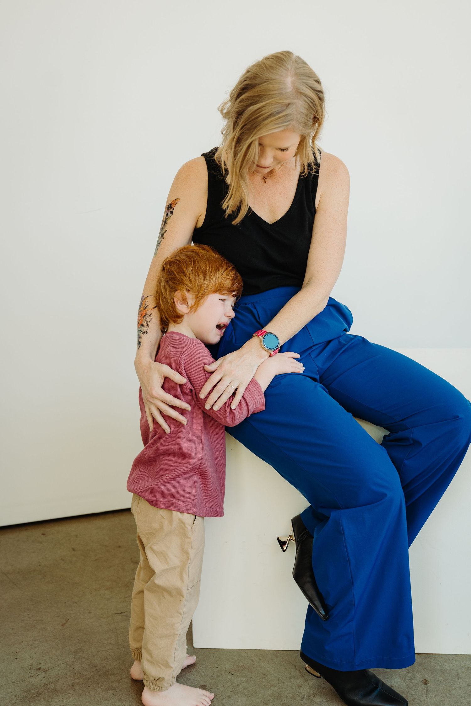 Blonde woman in blue flare pants sitting on white cube, hugging laughing red-haired boy in pink shirt.