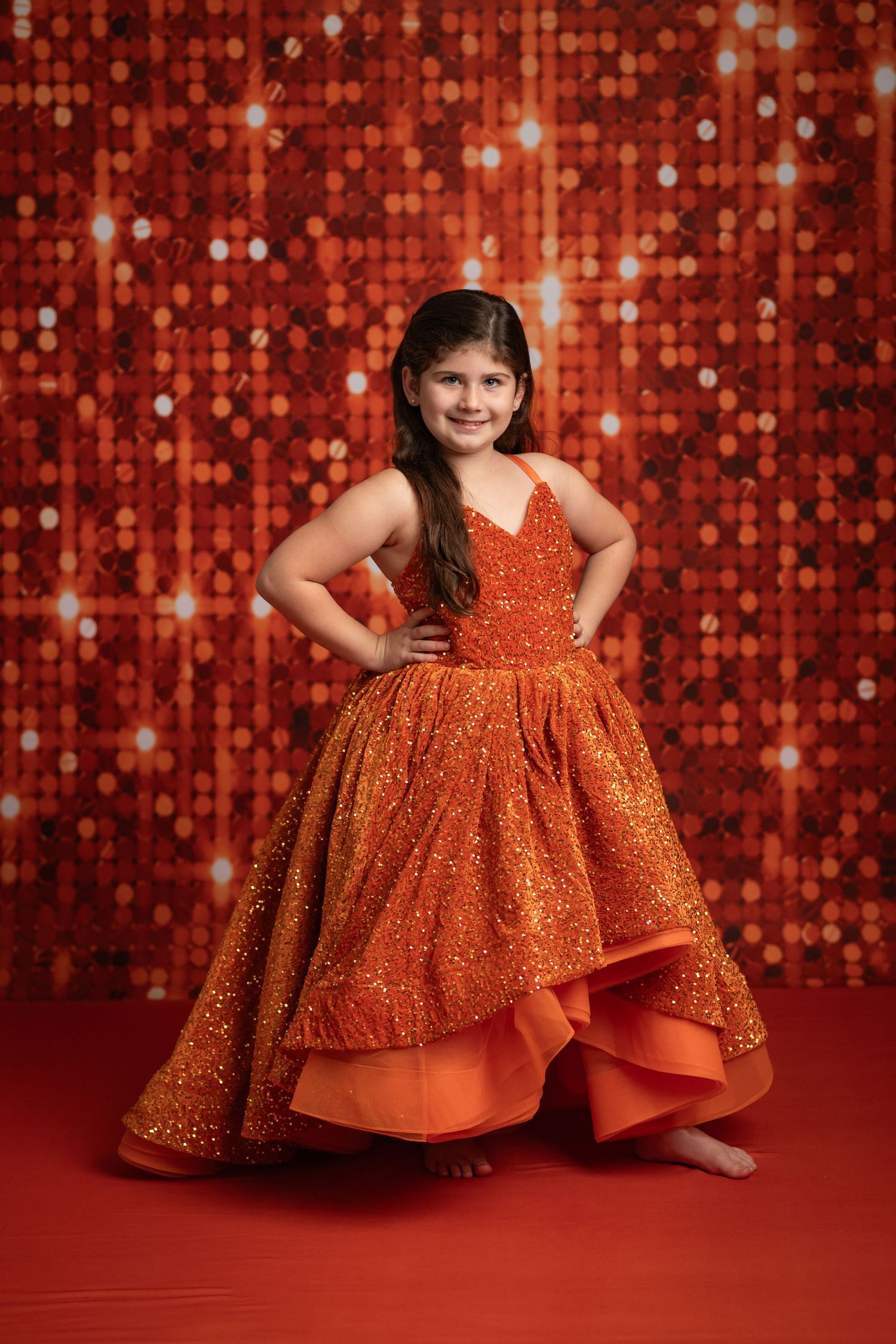 Young girl in sparkling orange ball gown poses confidently against a shimmering red sequin backdrop.