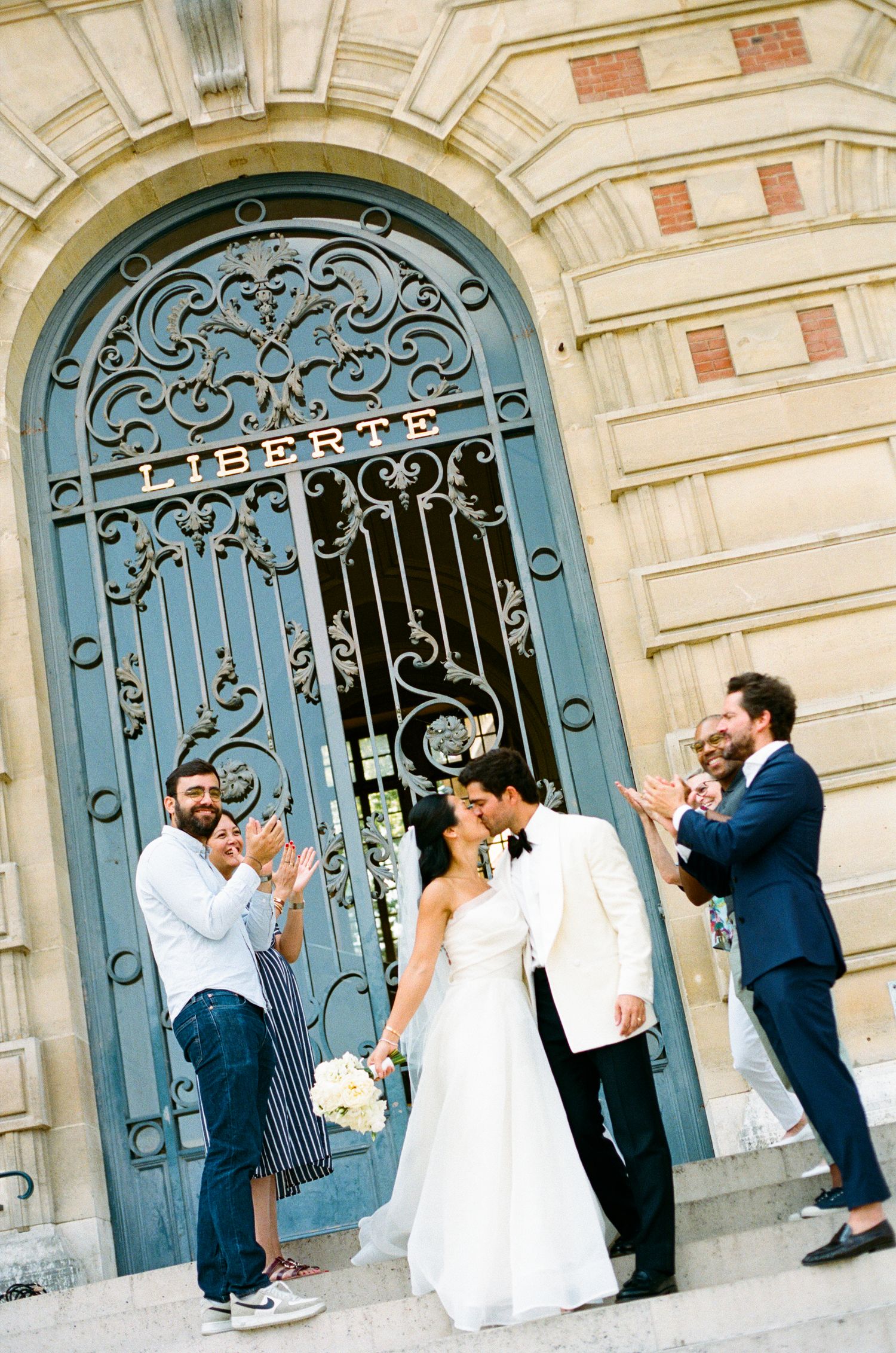 Bride and groom celebrate outside a Liberte gate in France, surrounded by clapping guests and a small dog. Versailles ceremony stairs