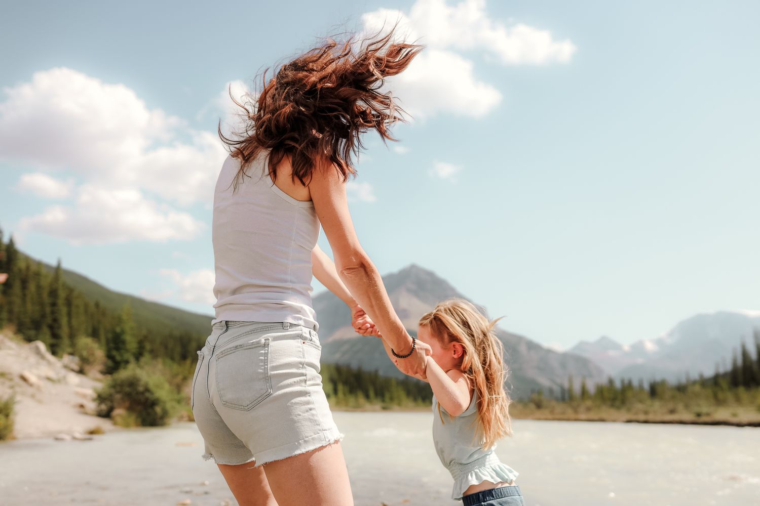 Mother and daughter dancing outdoors near a mountain river on a sunny day with blue skies.