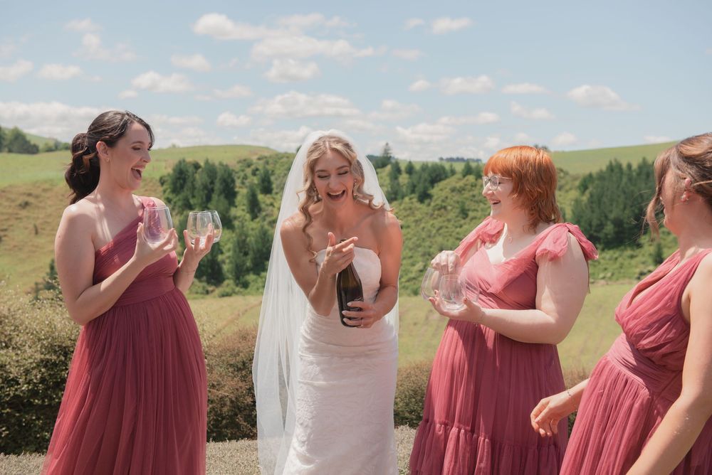 Bride in white gown opens champagne bottle with bridesmaids in dusty rose dresses outdoors on a sunny wedding day.