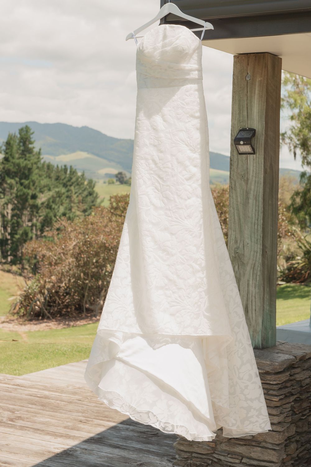 White lace wedding dress hanging on a hanger outdoors with scenic green hills and trees in the background.