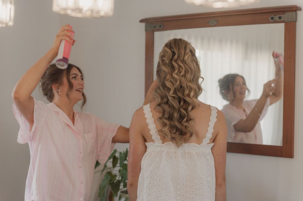 A bridesmaid sprays hairspray on a bride with long curly hair while she looks in a wooden-framed mirror.