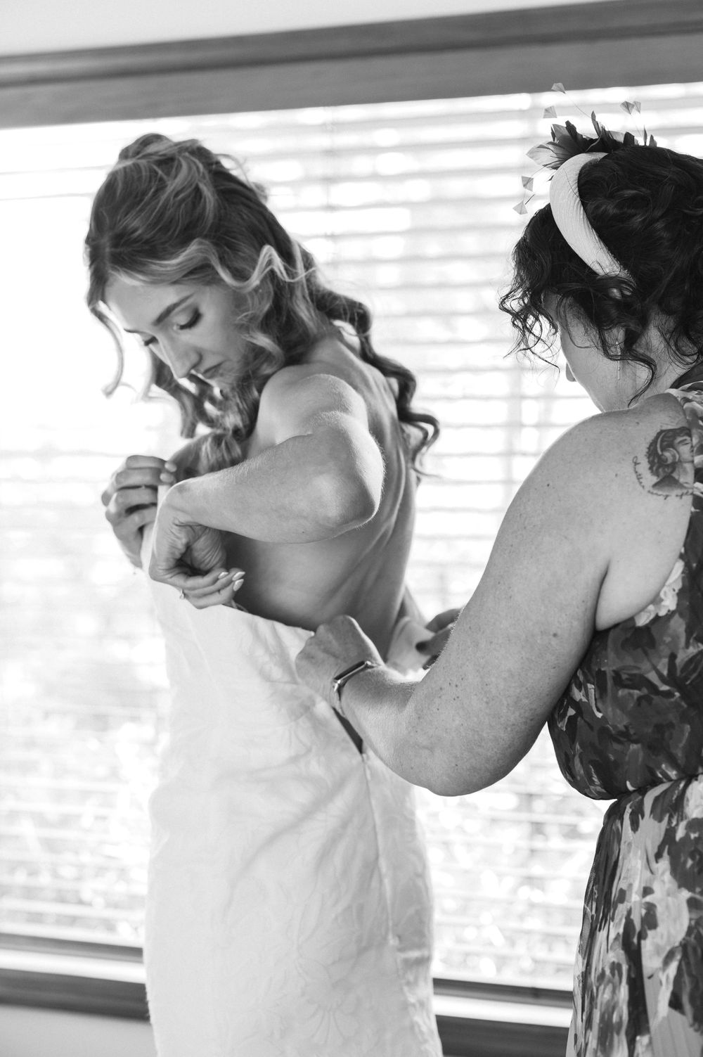 Black and white photo of a bride getting her wedding dress fastened by a bridesmaid near a window.