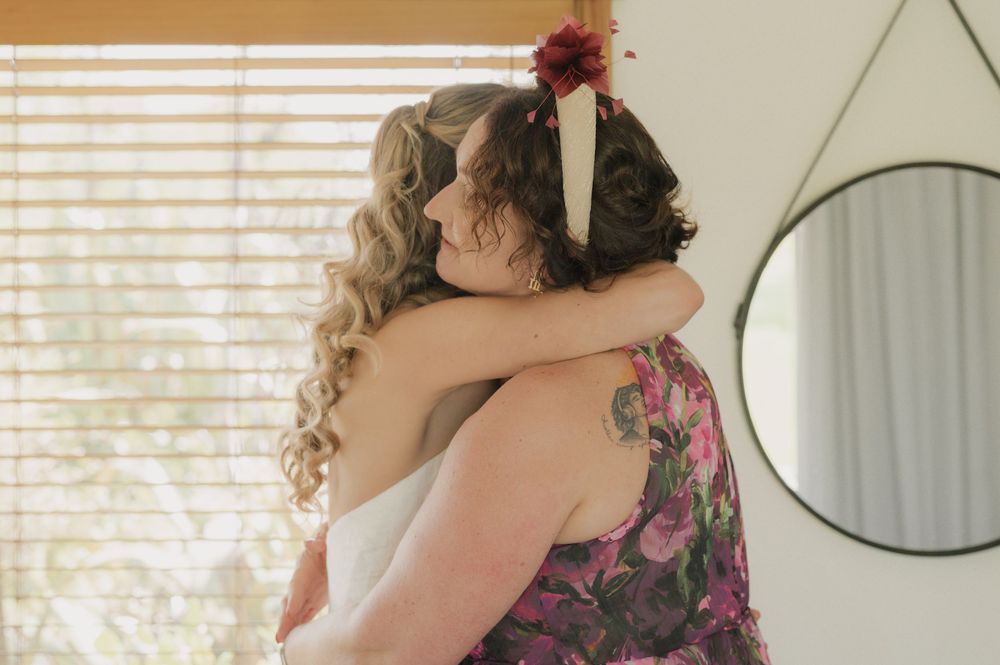 Two women share a heartfelt hug in a bright room, one in a white wedding dress and the other in a floral dress.
