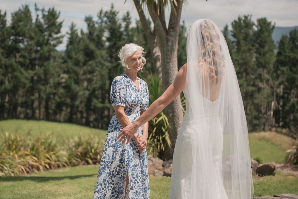Bride in white gown and veil holds hands with elderly woman in floral dress outdoors at a garden wedding.
