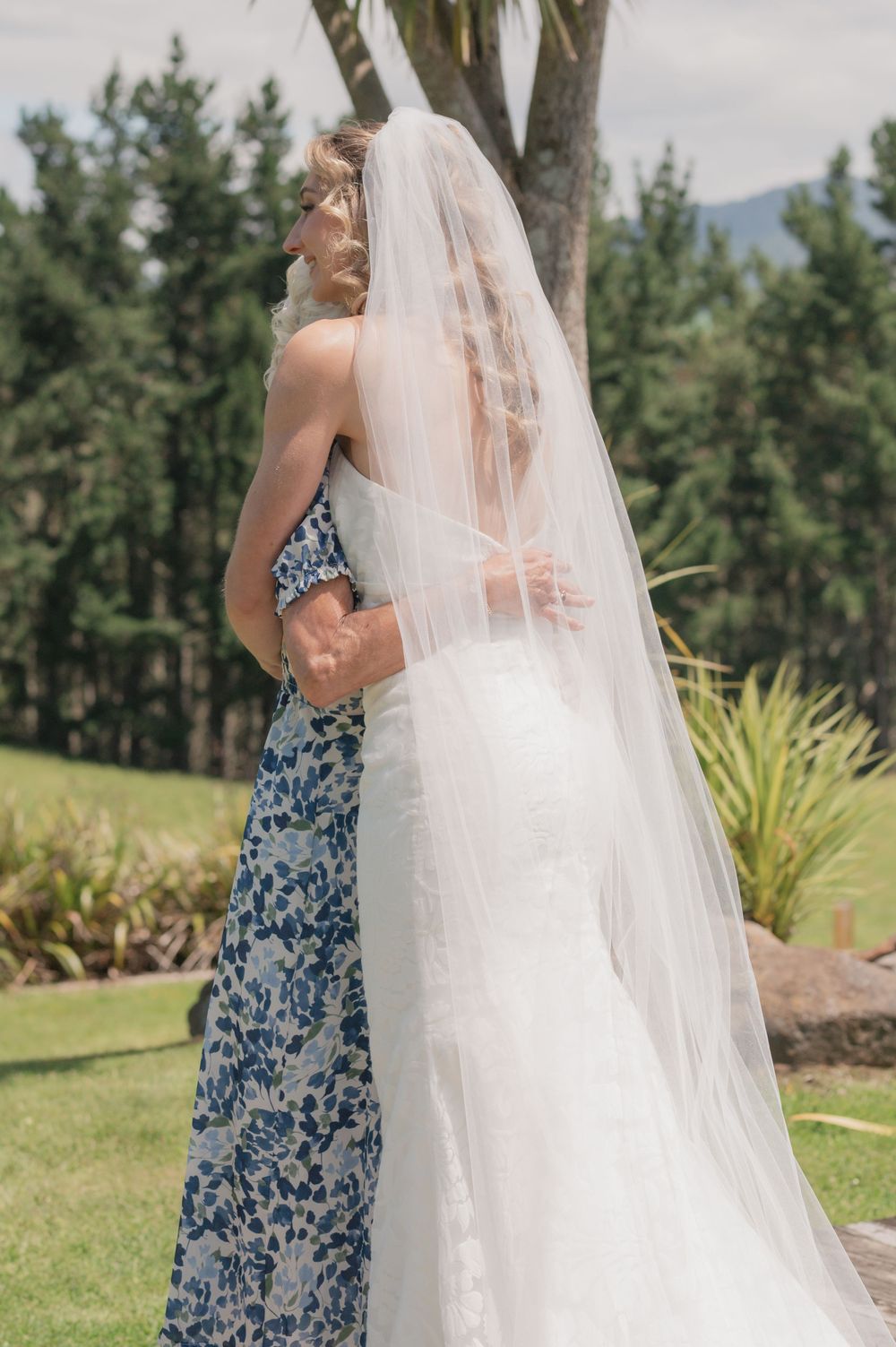 Bride in white gown and veil embracing woman in blue floral dress outdoors at wedding ceremony.