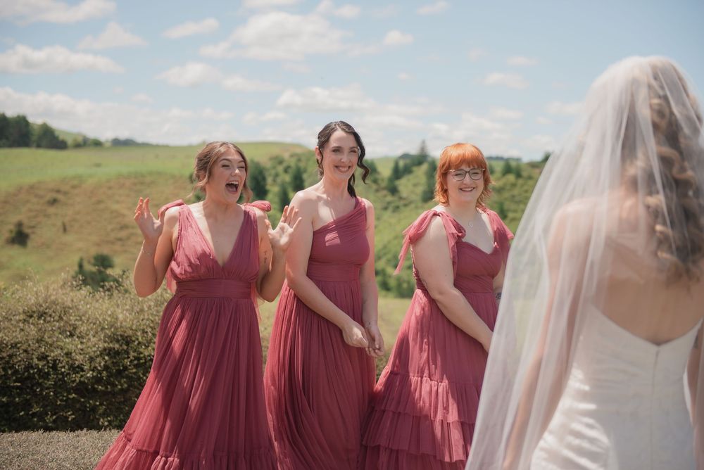 Three bridesmaids in dusty rose dresses react with joy and excitement seeing the bride in her white wedding gown outdoors.