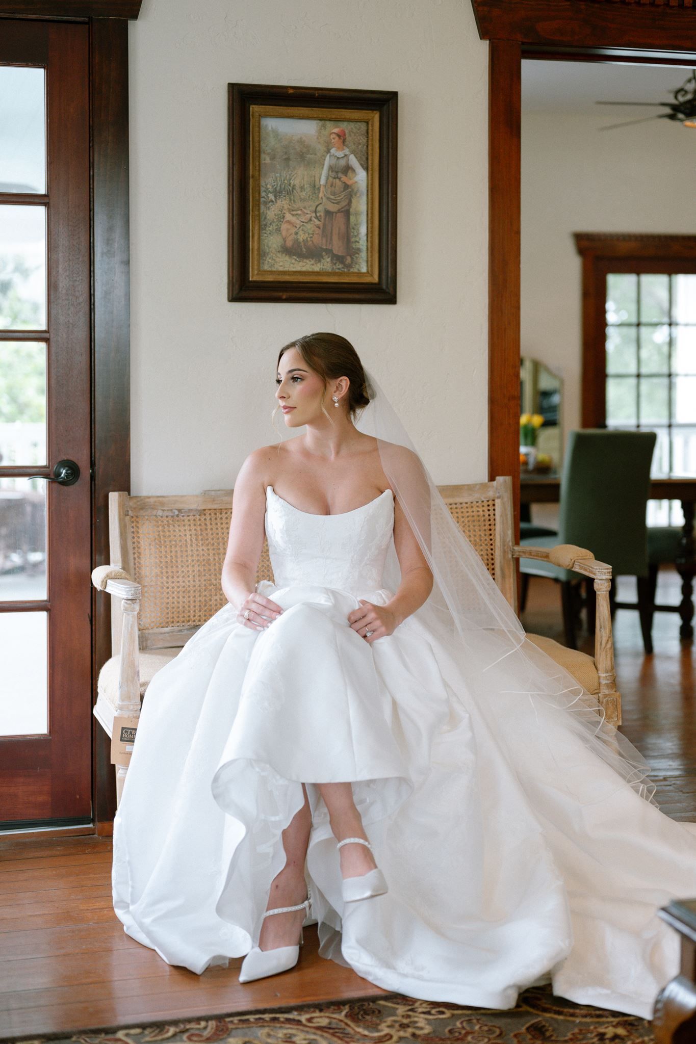 Bride in strapless white ball gown with veil sits gracefully on cane chair in elegant room with wood doors.