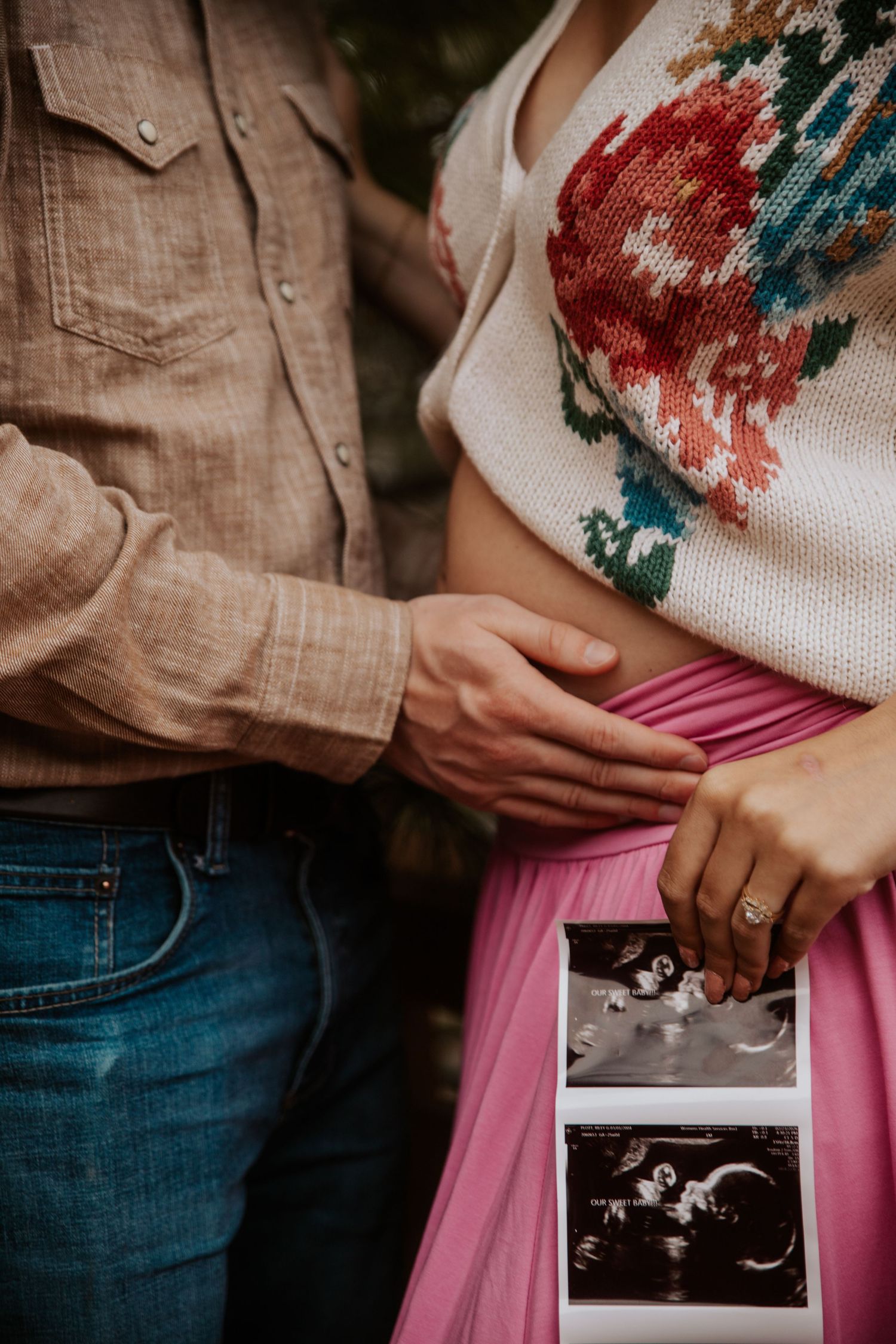 Pregnant couple holding ultrasound photos, woman in floral sweater with pink skirt, man in beige shirt and jeans touching baby bump.