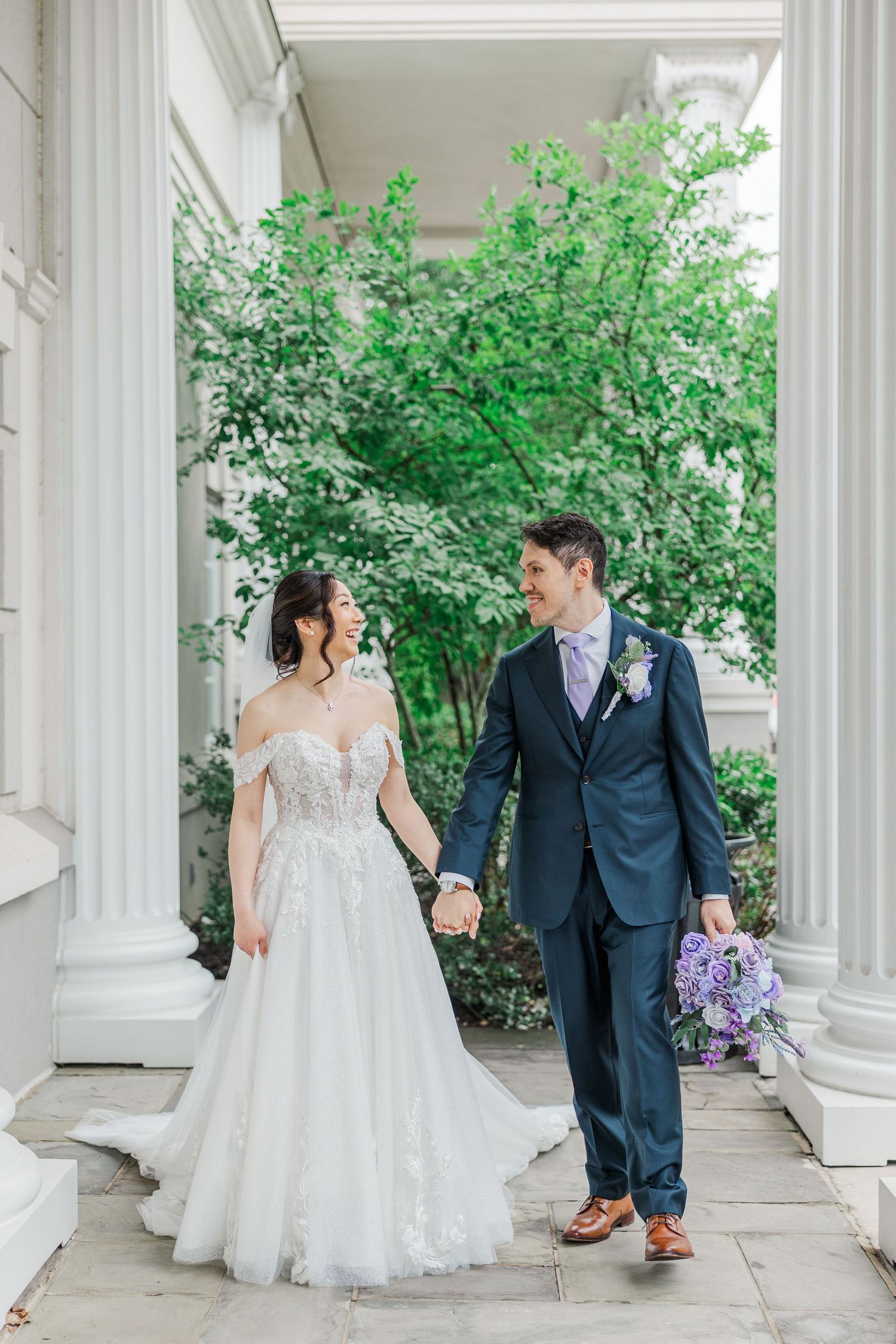Bride and groom walk hand-in-hand between white columns, she in lace gown, he in navy suit with purple boutonniere.
