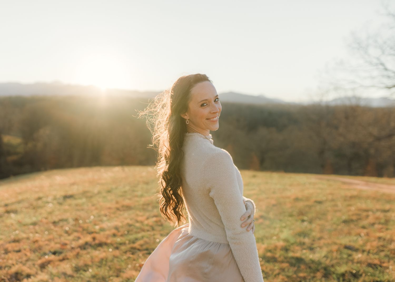 Woman smiling over shoulder at golden hour in a sunlit field with rolling hills and warm autumn light.
