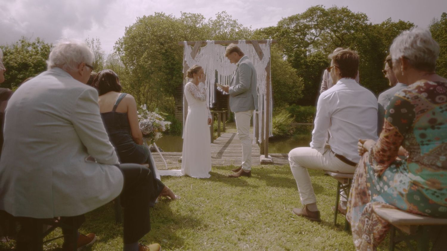 Outdoor wedding ceremony with couple exchanging vows under white arch, guests seated on chairs in lush green garden.