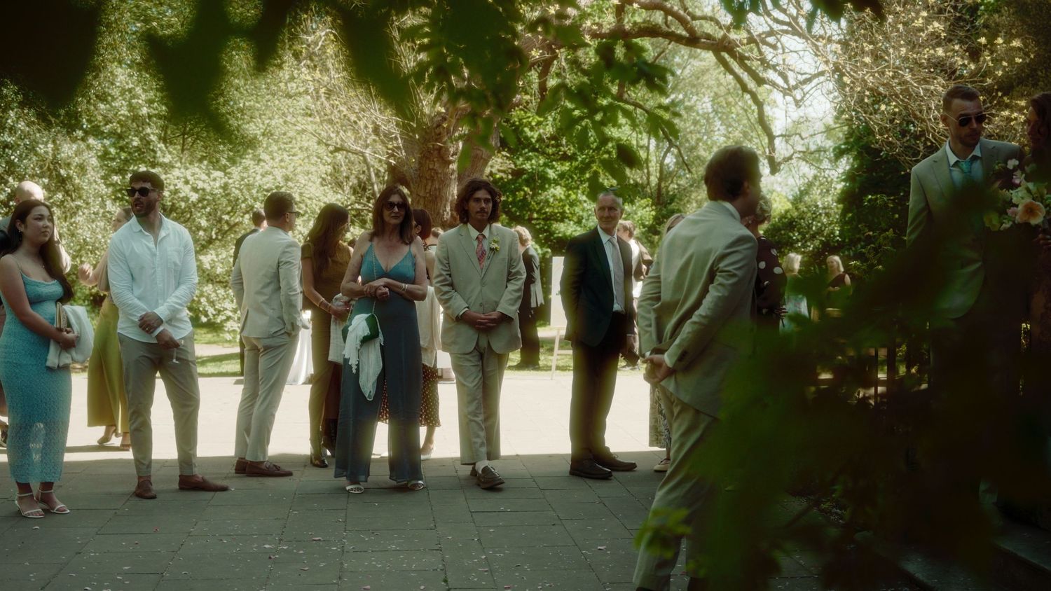 Elegantly dressed wedding guests mingle outdoors in a lush green garden setting during a summer ceremony.