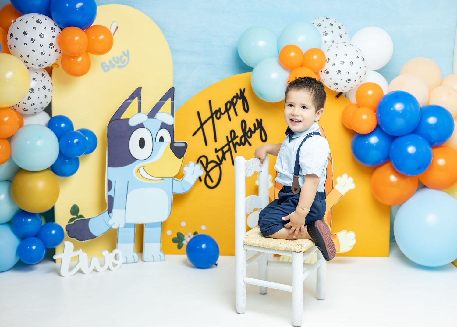 Toddler boy sits on chair at Bluey themed birthday party with colorful balloons and Happy Birthday backdrop.