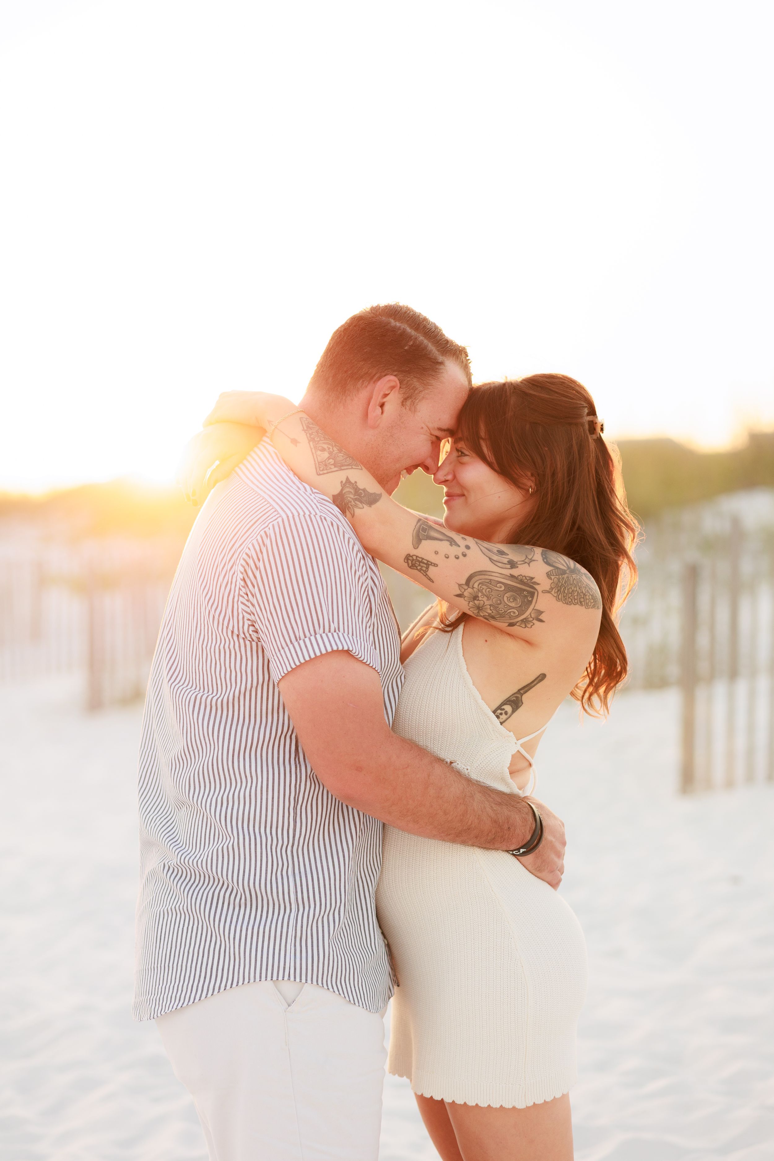 Couple embracing at sunset on a sandy beach, woman in white dress with tattoos, golden backlight.