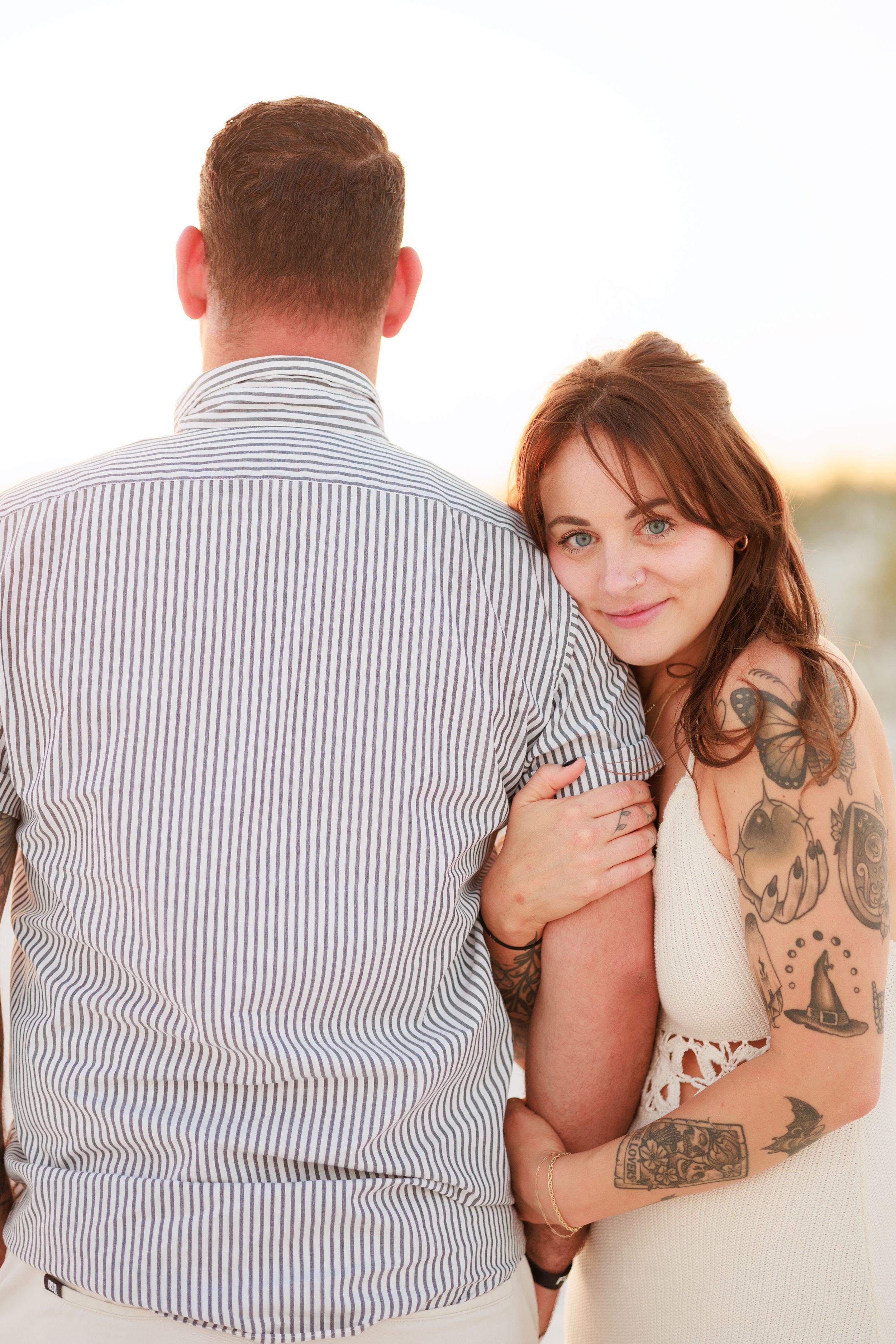 A tattooed woman smiles over her shoulder while embracing a man in a striped shirt outdoors.